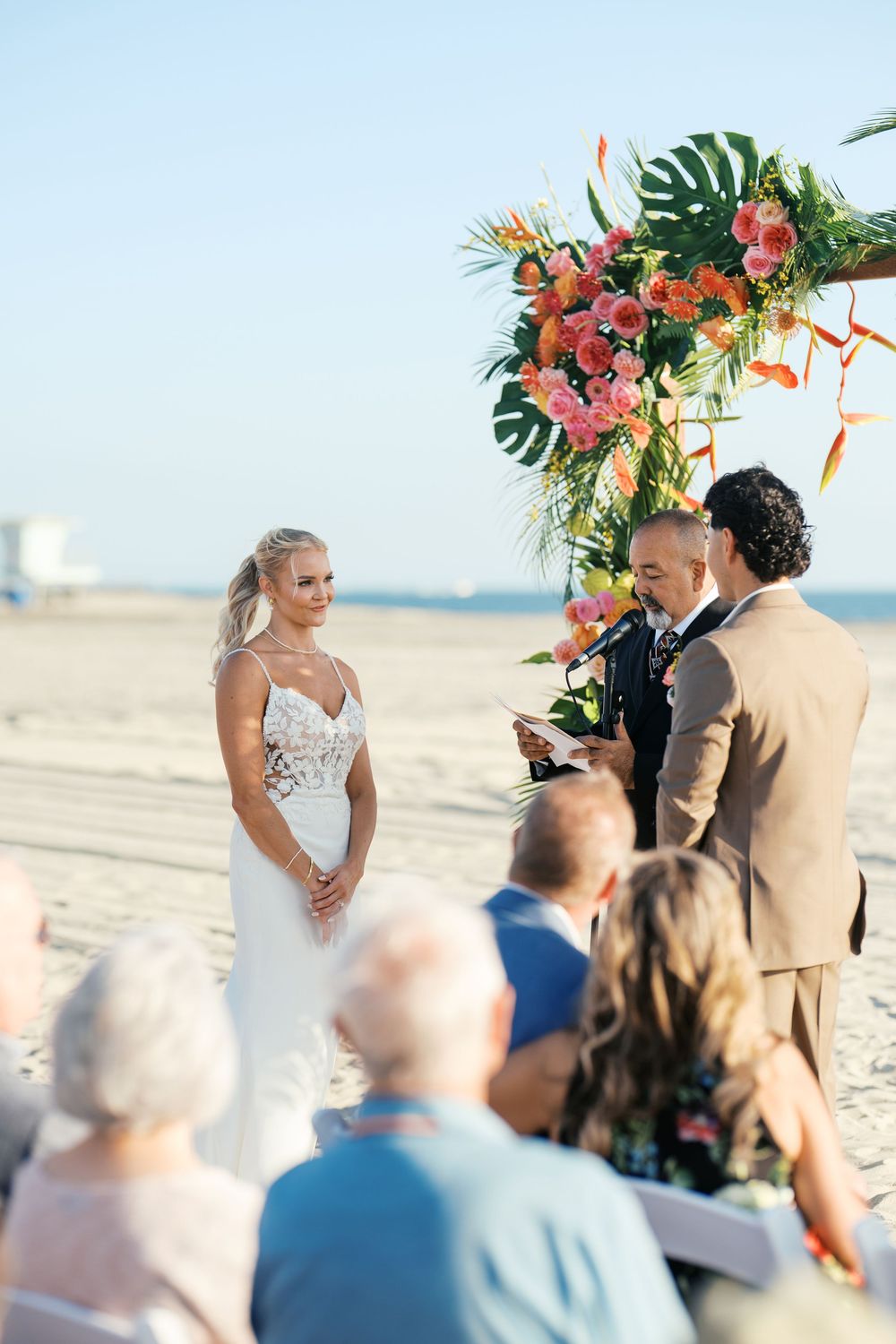 A beach wedding ceremony takes place under a tropical floral arch with pink flowers and monstera leaves as guests look on.