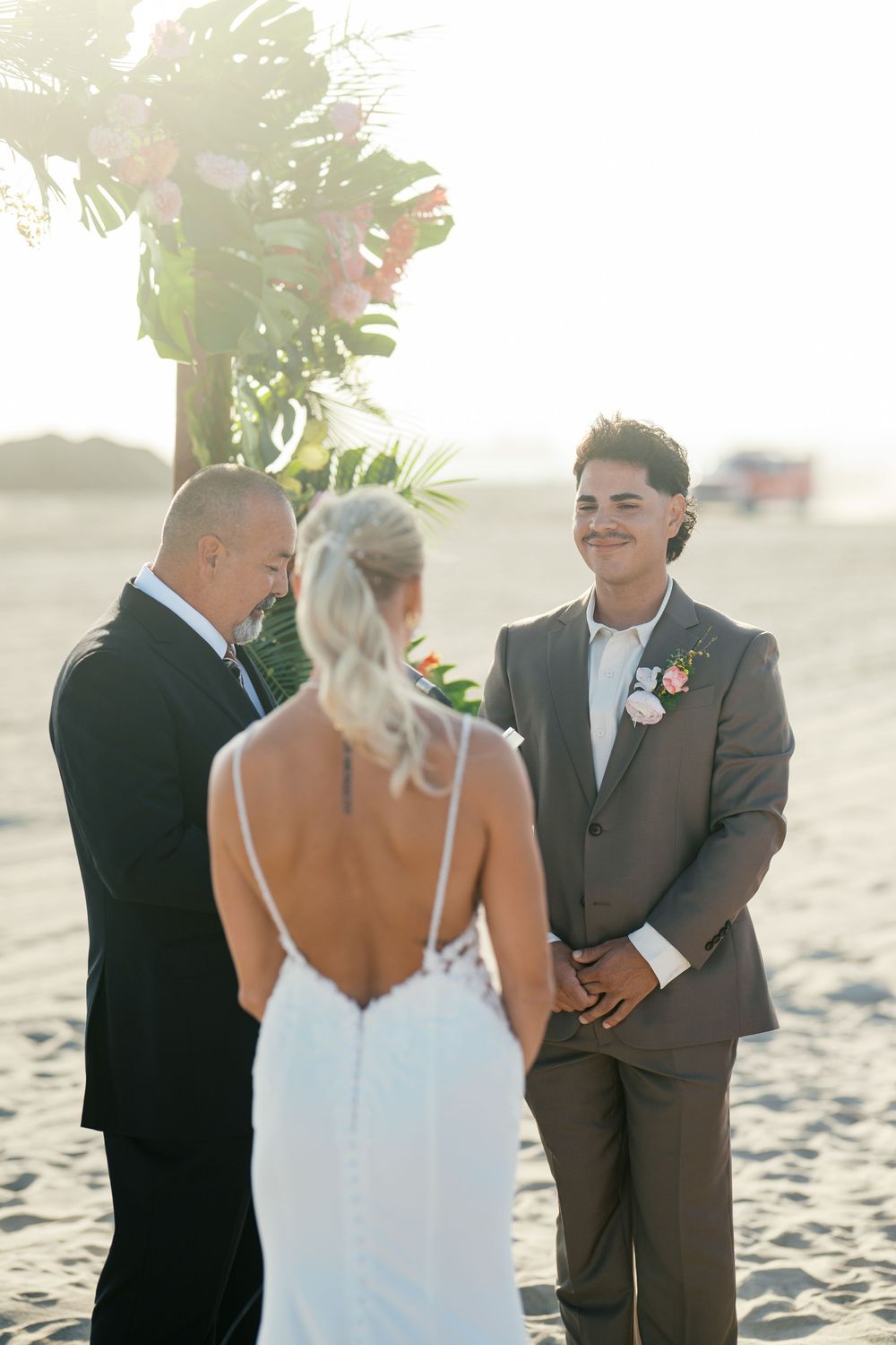 A beachside wedding ceremony takes place at sunset with palm trees and warm lighting in the background.