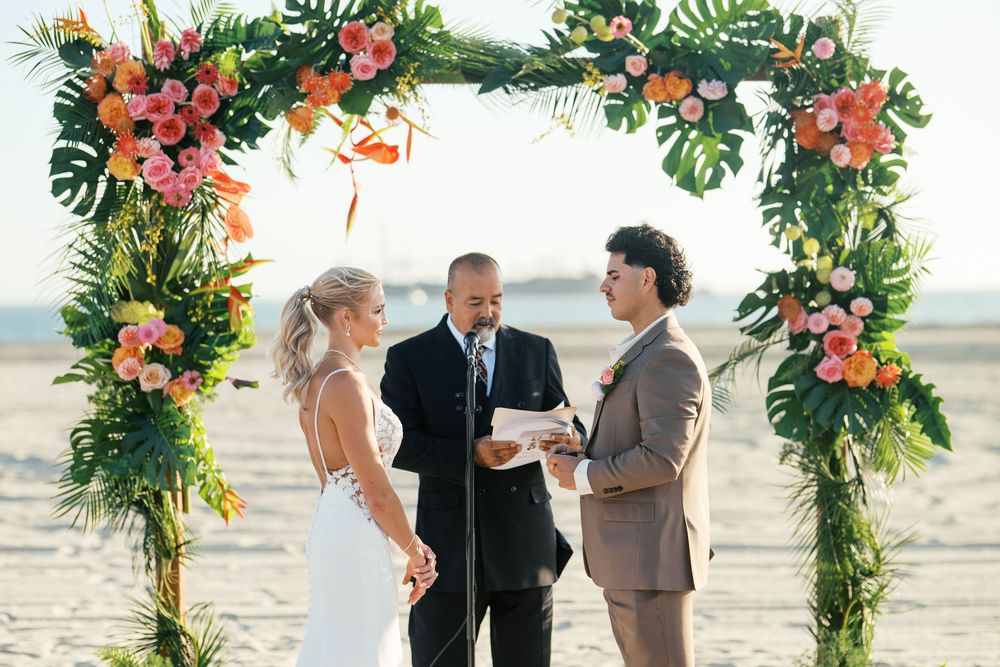 Happy couple exchanges vows during beach wedding ceremony beneath tropical floral arch with pink blooms and greenery.