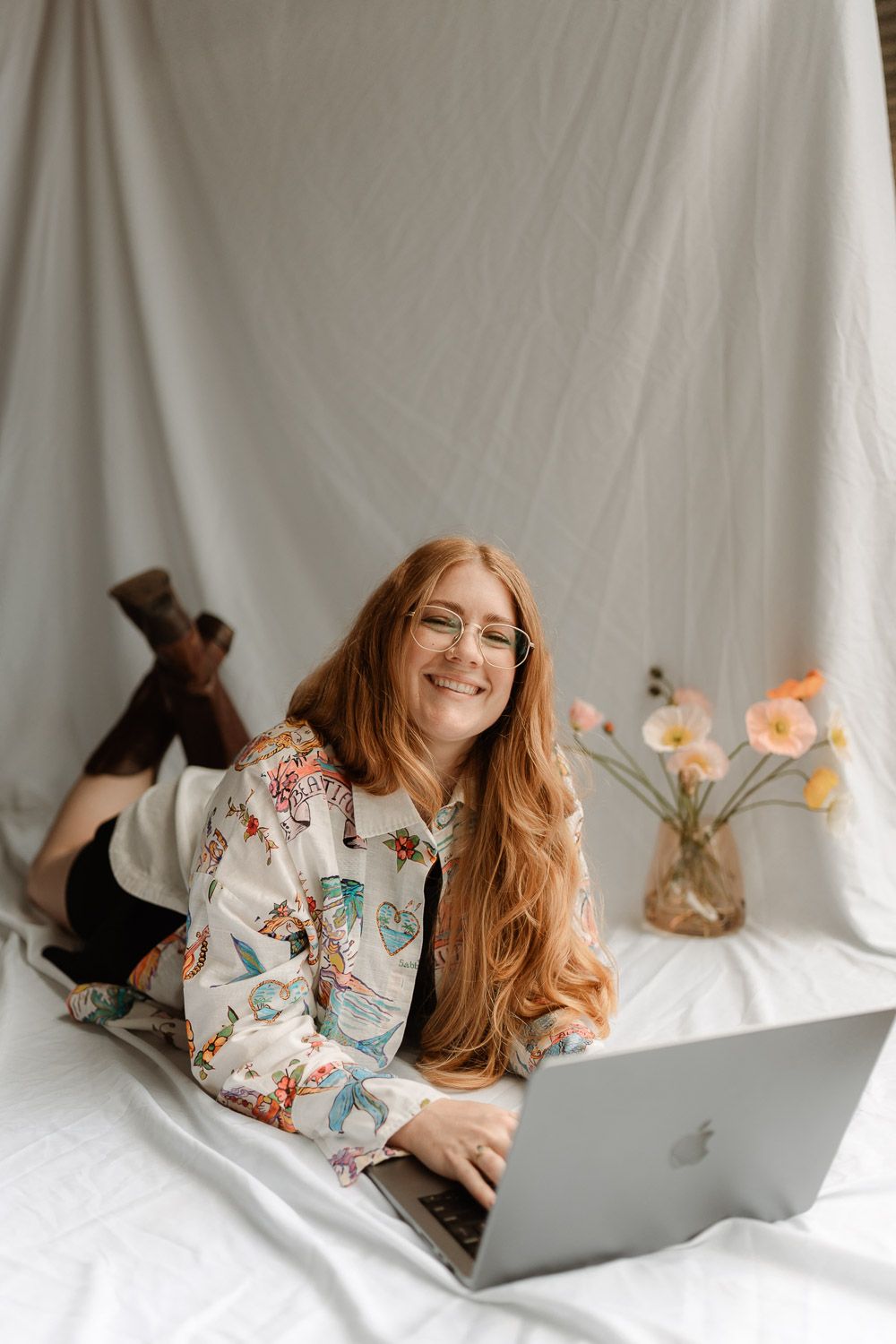 Person in butterfly print pajamas working on laptop while lying on white bedding in bright natural light.