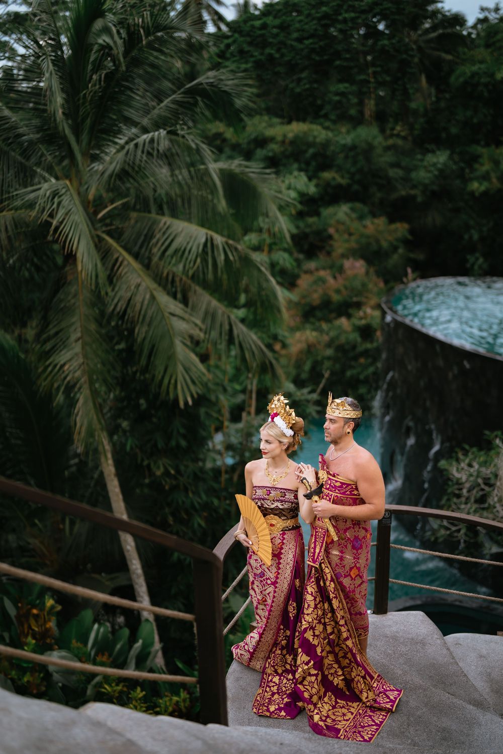 Couple in traditional Balinese dress pose near waterfall and palm trees.