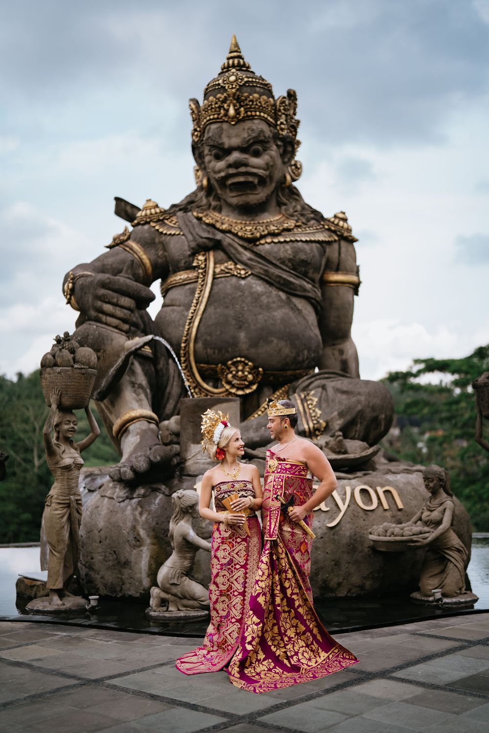 Couple in ceremonial dress pose before large Balinese temple guardian statues.