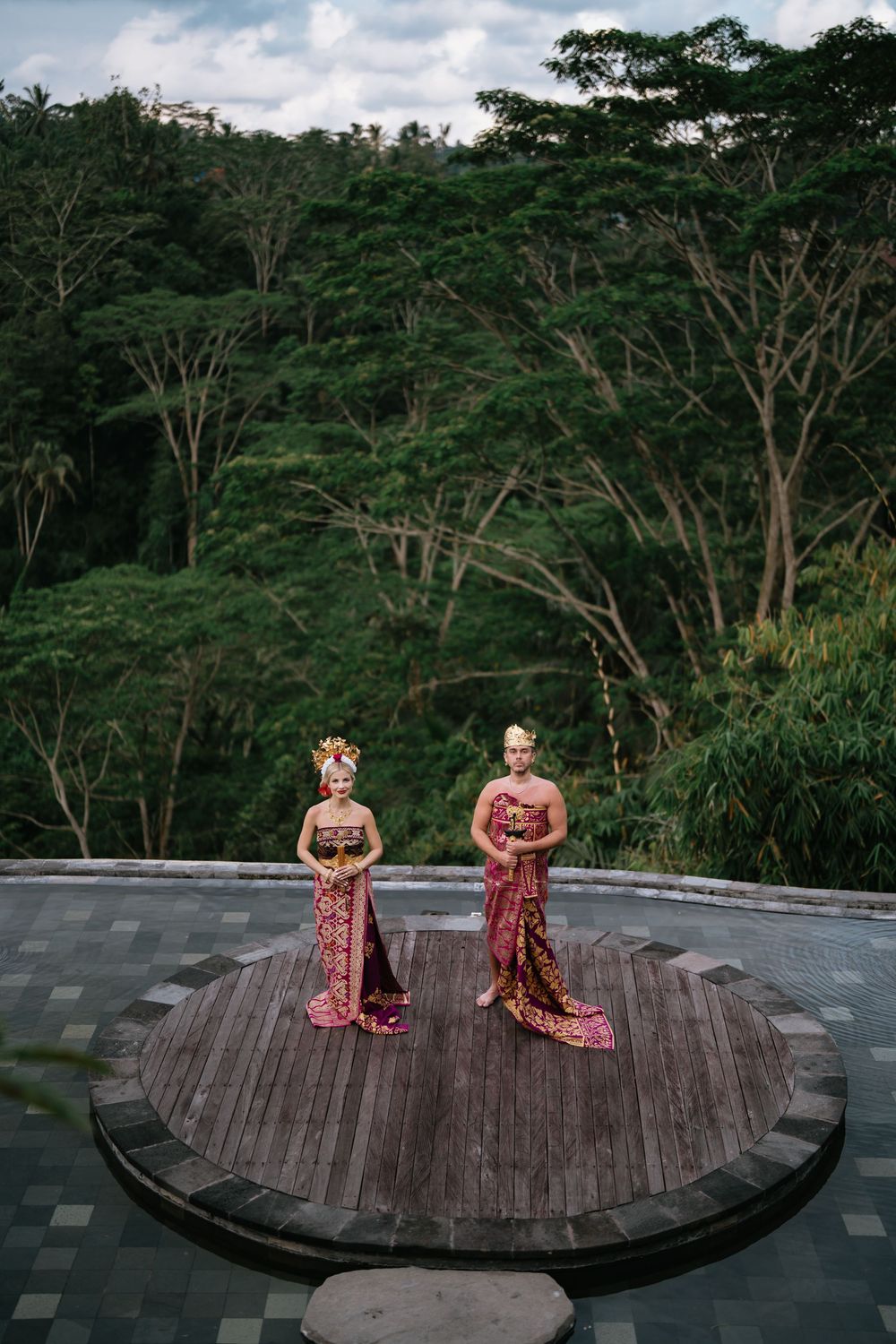 Couple in traditional dress photographed on circular wooden platform surrounded by forest.