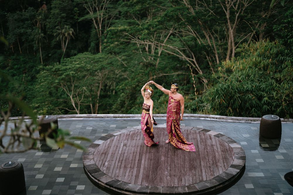 Two figures in traditional Balinese dress perform a cultural dance on a wooden deck surrounded by lush greenery.