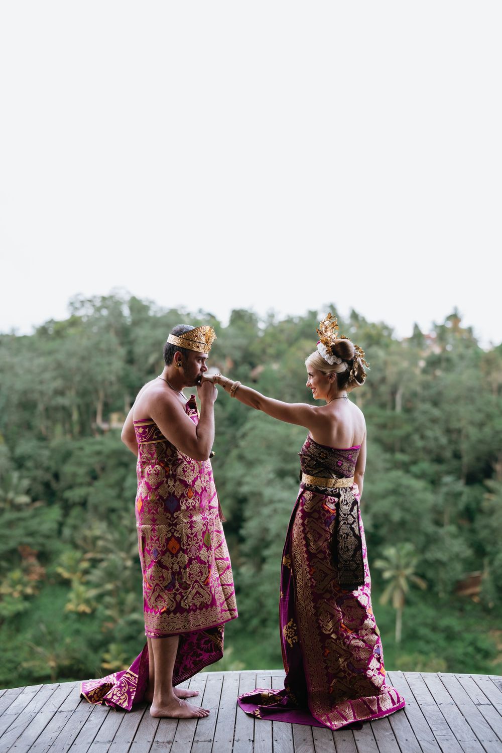 Traditional dancers wearing pink and burgundy Balinese costumes sit on a wooden platform with tropical forest backdrop.