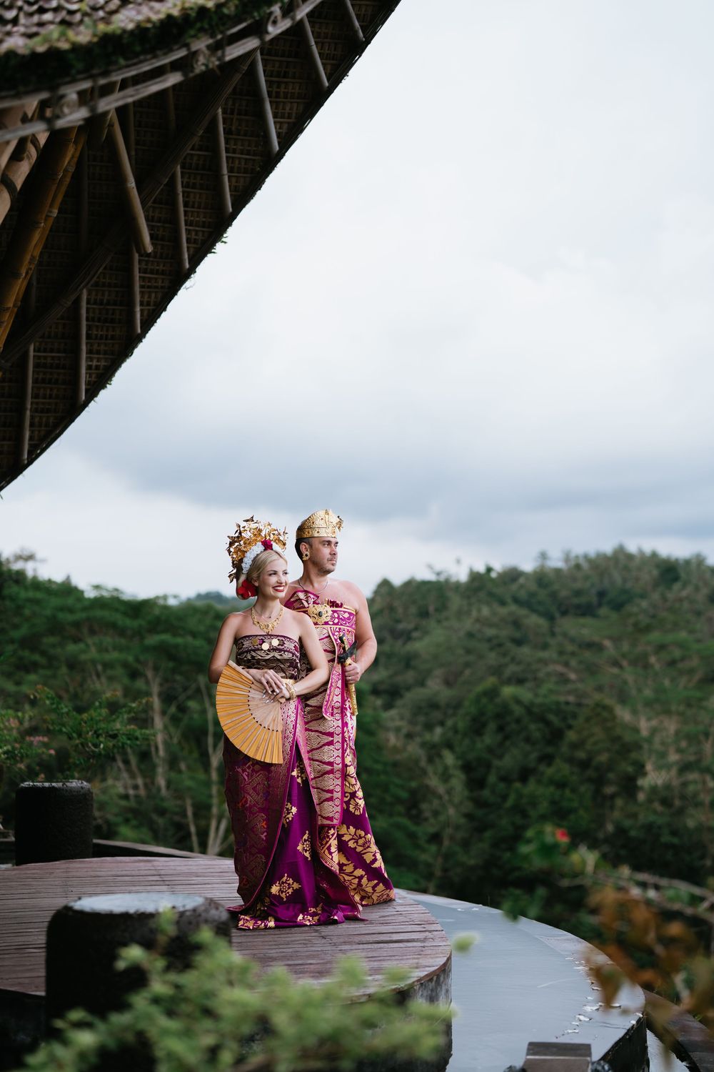 Two people in traditional Balinese dress sit together on wooden platform with mountain backdrop.