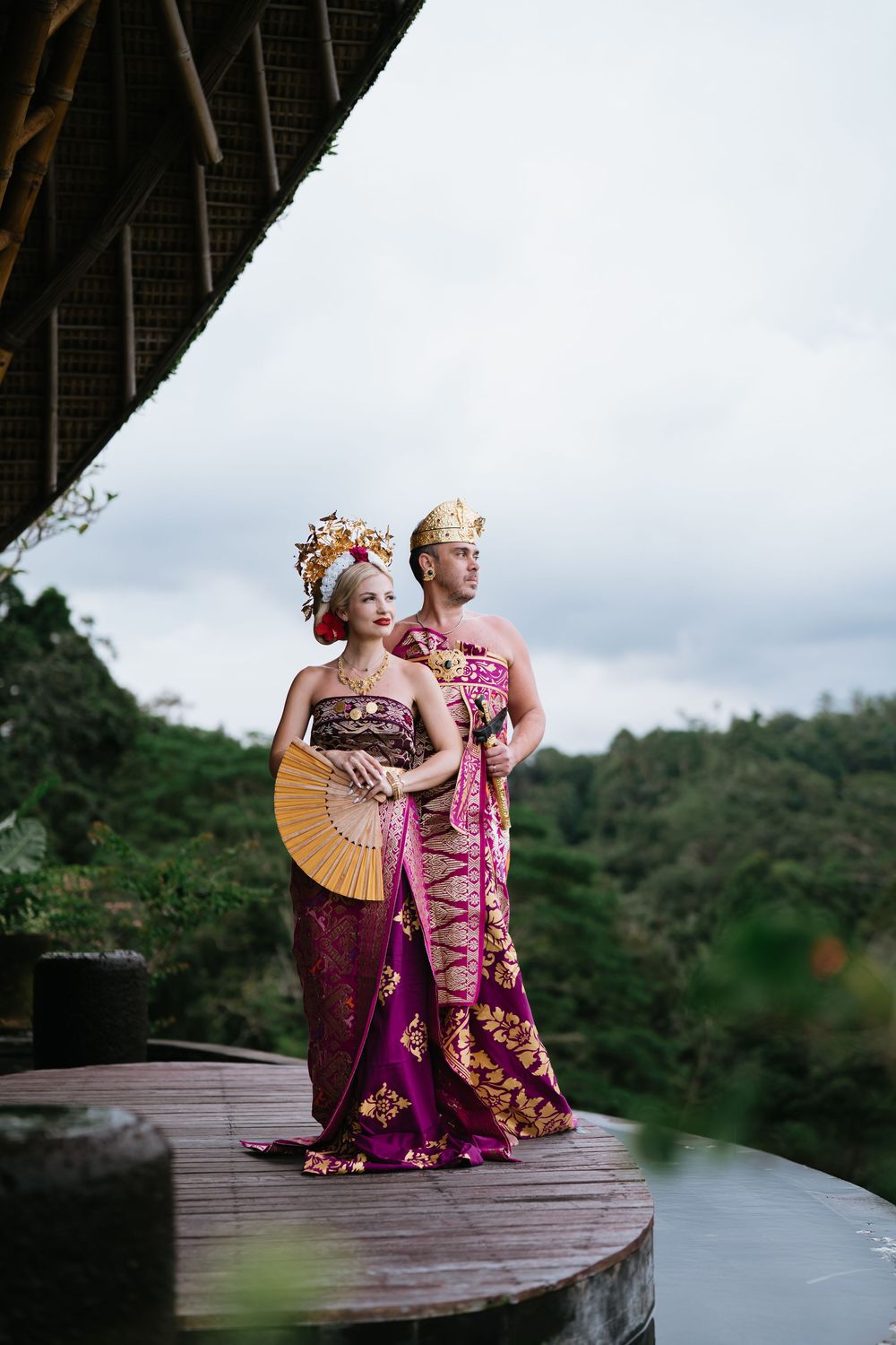 Couple in ornate Balinese costumes pose with traditional fan against cloudy sky.