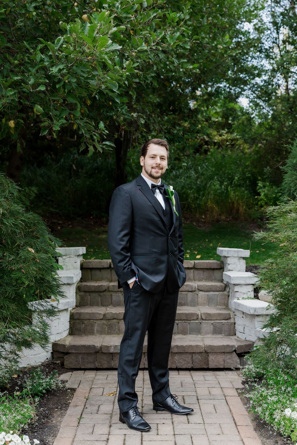 Groom in classic black tuxedo poses on brick steps in garden setting at The Wren House in St. Jacobs, Ontario.