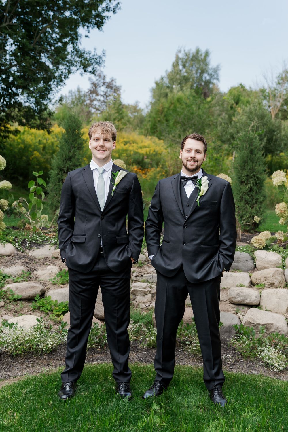 Two groomsmen in formal attire pose casually in garden setting at The Wren House in St. Jacobs, Ontario.