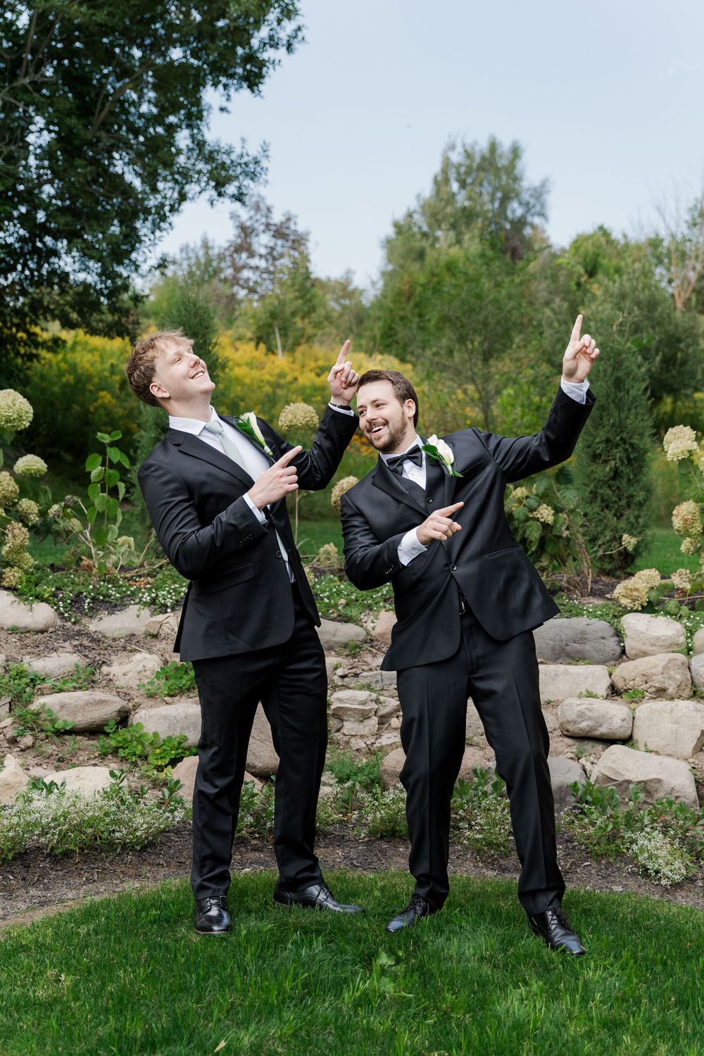 Groomsmen in black tuxedos strike playful poses in garden setting at The Wren House in St. Jacobs, Ontario.