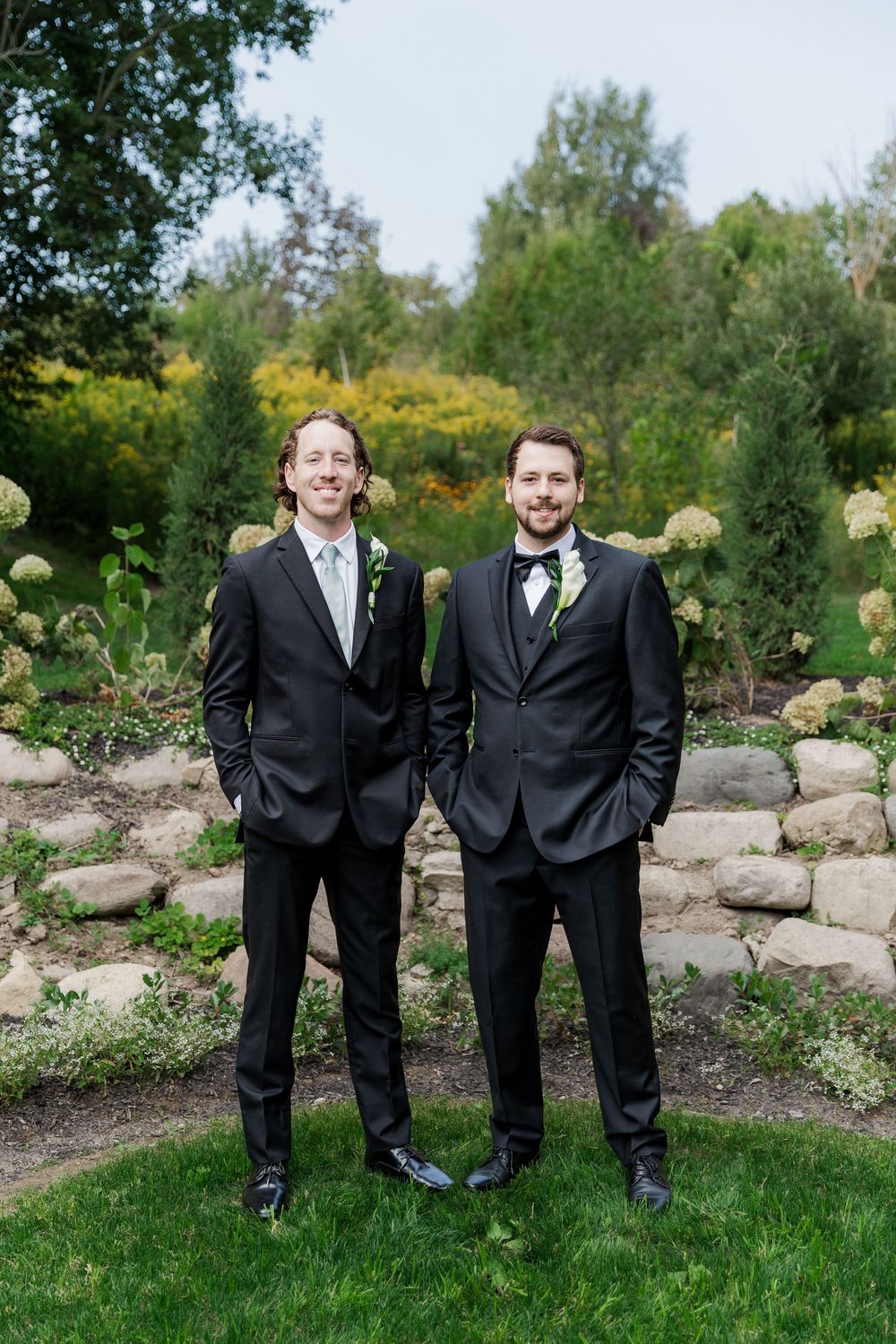 Two groomsmen in black suits stand together on lawn with stone wall backdrop at The Wren House in St. Jacobs, Ontario.
