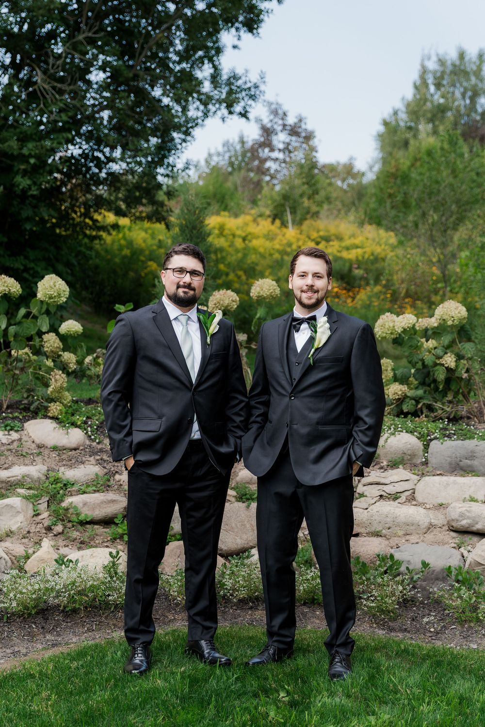 Groomsmen in black tuxedos pose together in a garden setting with white hydrangeas and rocks at The Wren House in St. Jacobs, Ontario.