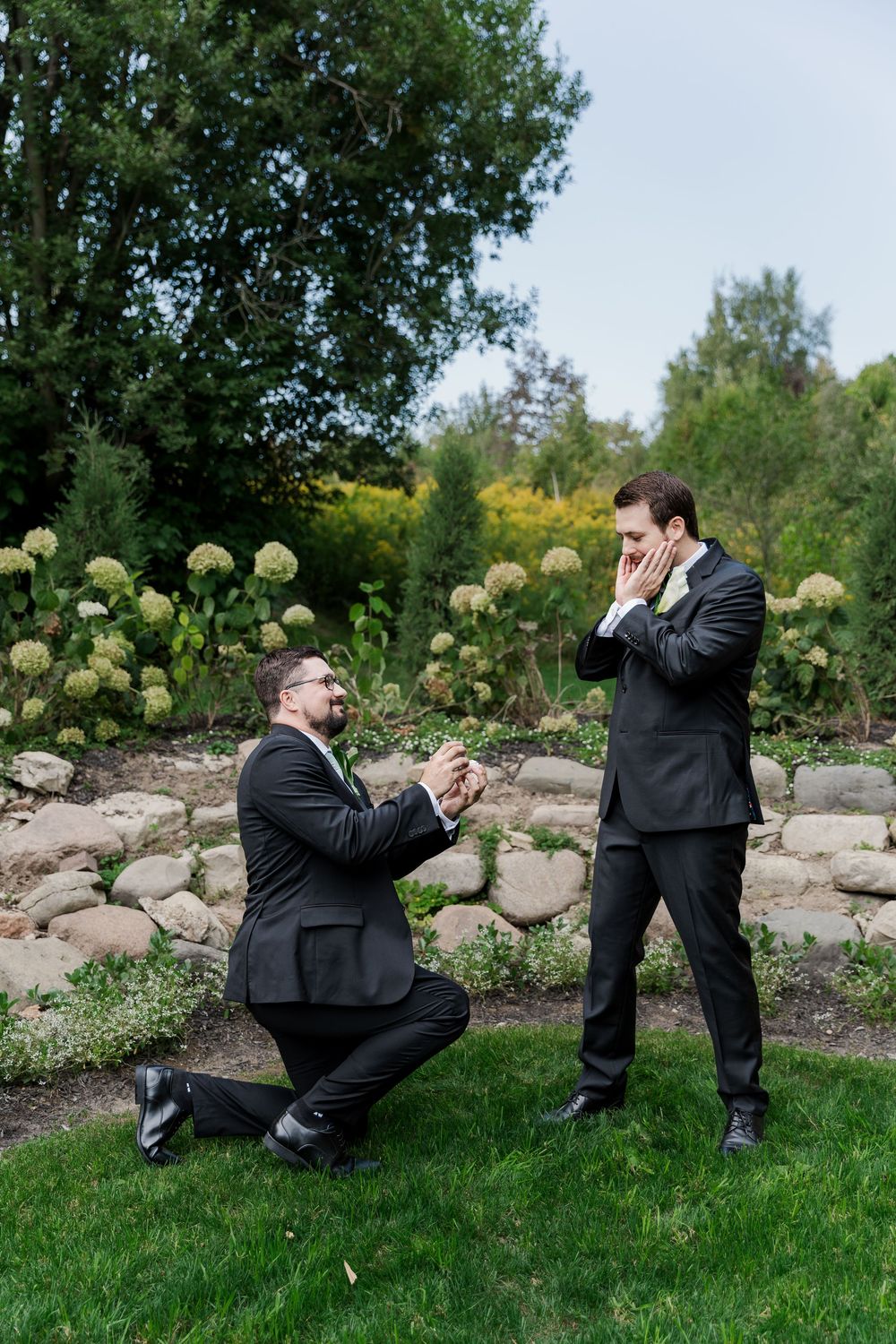 Wedding party members in formal attire playfully pose in a landscaped garden setting at The Wren House in St. Jacobs, Ontario.
