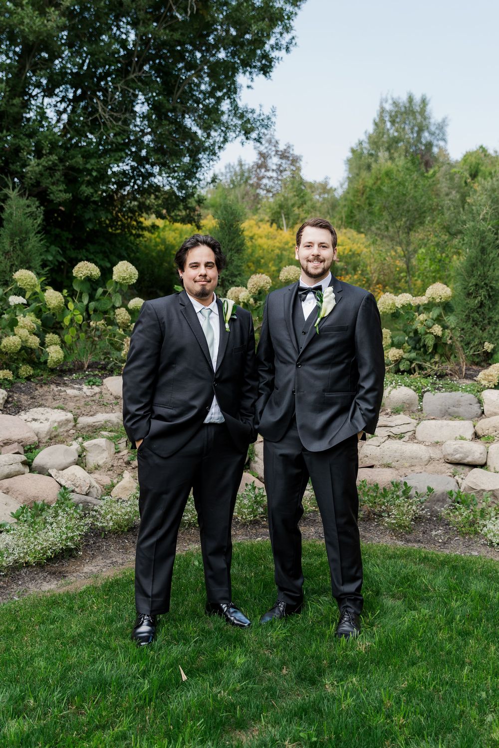 Groomsmen share a candid moment while standing on grass with decorative rocks and flowers at The Wren House in St. Jacobs, Ontario.