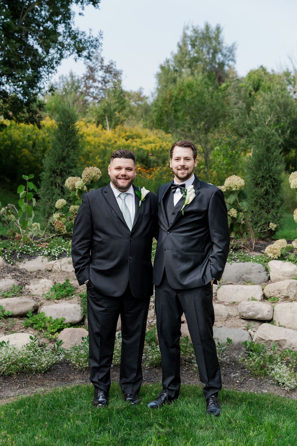 Groomsmen in matching black tuxedos stand together on grass with a landscaped garden backdrop at The Wren House in St. Jacobs, Ontario.