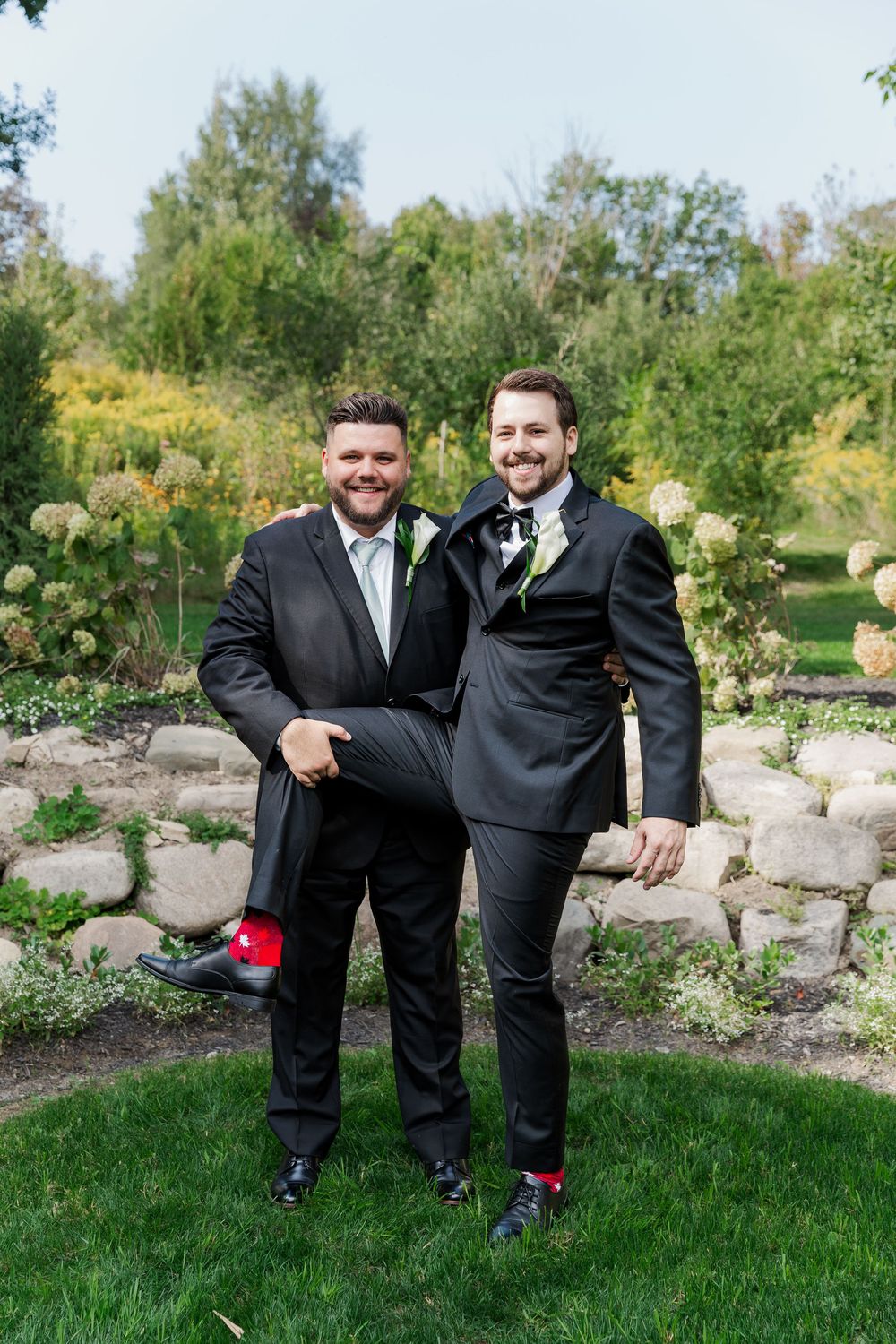 Groomsmen in black suits strike playful poses on grass with decorative garden rocks behind them at The Wren House in St. Jacobs, Ontario.
