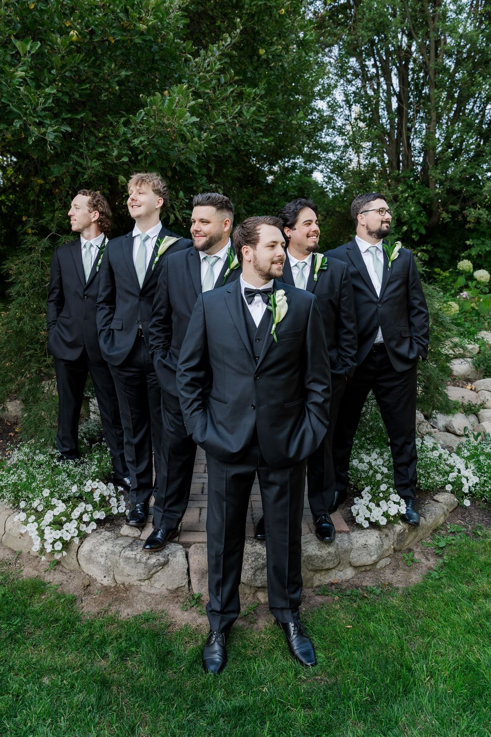Wedding party arranged in casual poses against a natural stone wall and garden backdrop at The Wren House in St. Jacobs, Ontario.