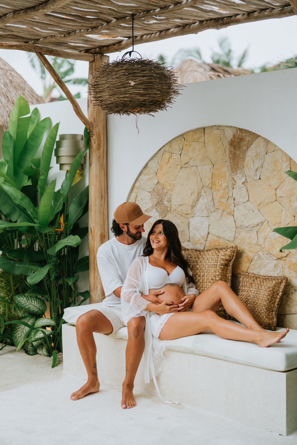Couple poses intimately on white daybed with tropical decor and plants.
