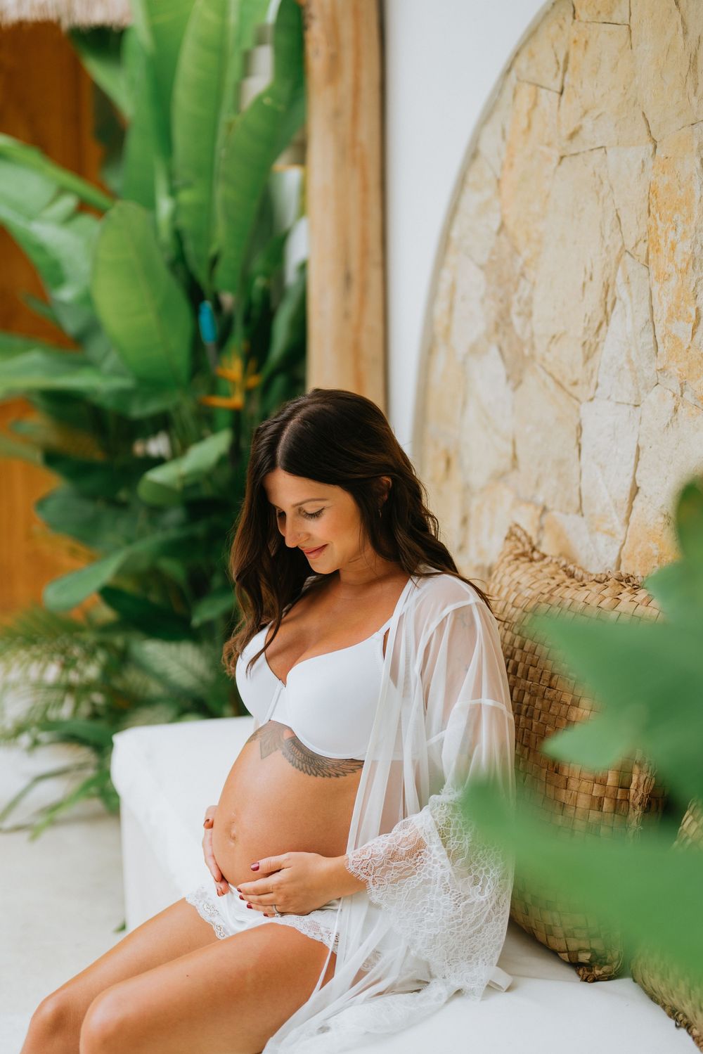 Maternity photo shoot in white lace robe with tropical plants in background.