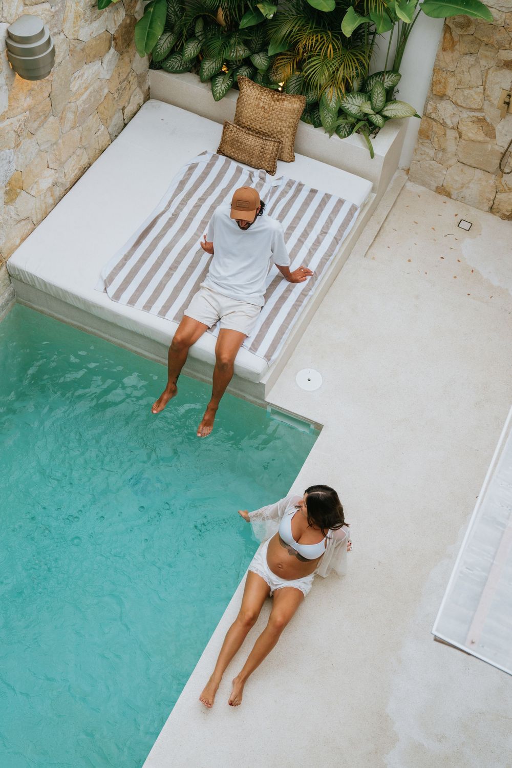 Couple sitting on edge of modern pool deck with tropical garden surroundings.