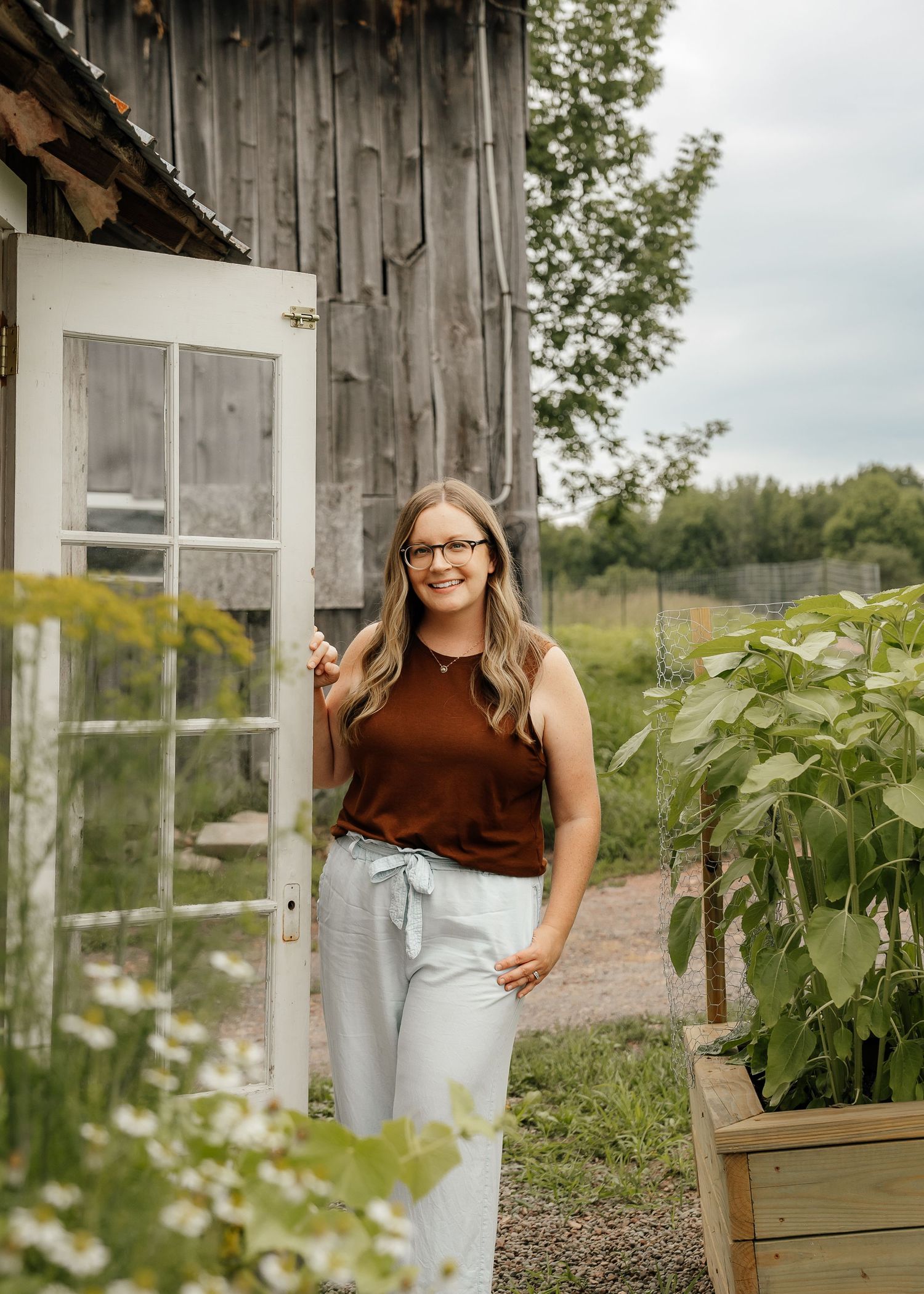 Person in brown top and light pants stands near wooden greenhouse surrounded by lush garden plants and sunflowers.