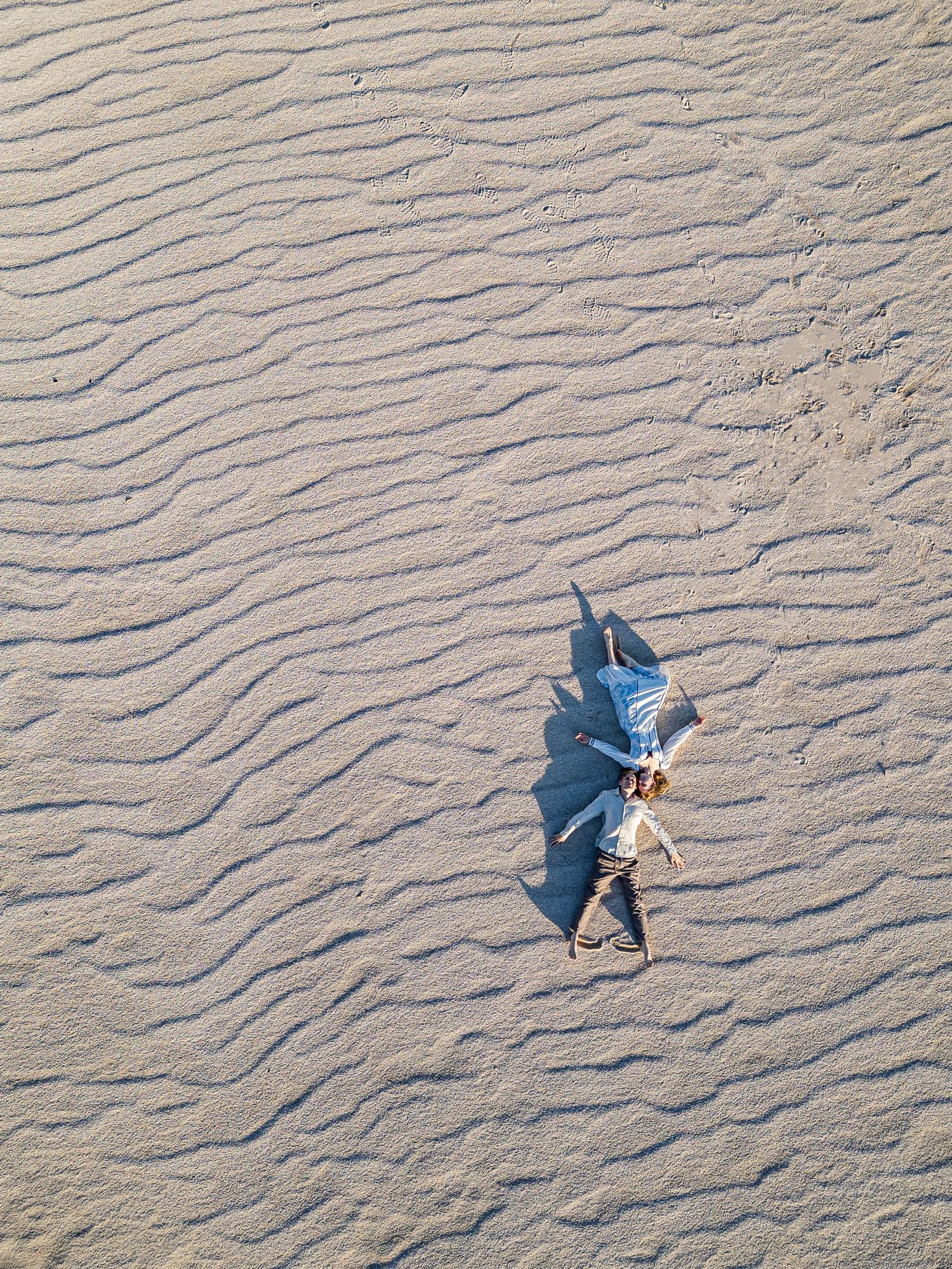 Two people cast long shadows while walking on a textured sandy beach with ripple patterns at sunset.