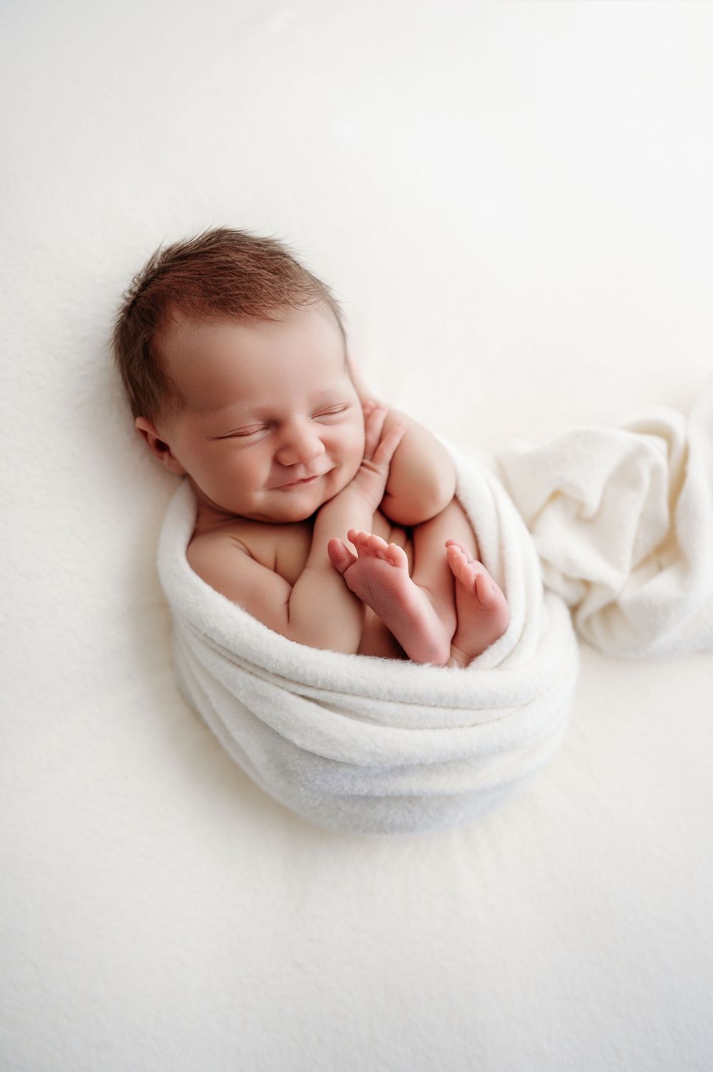 Peaceful sleeping newborn baby wrapped in soft white blanket against light background.