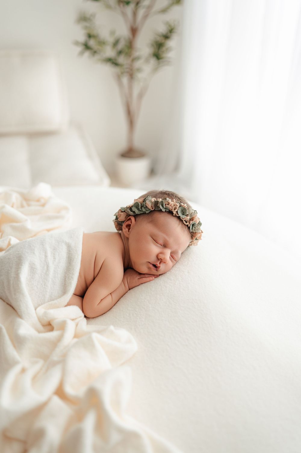 Peaceful sleeping newborn baby wearing floral crown on white blanket with soft natural window light.