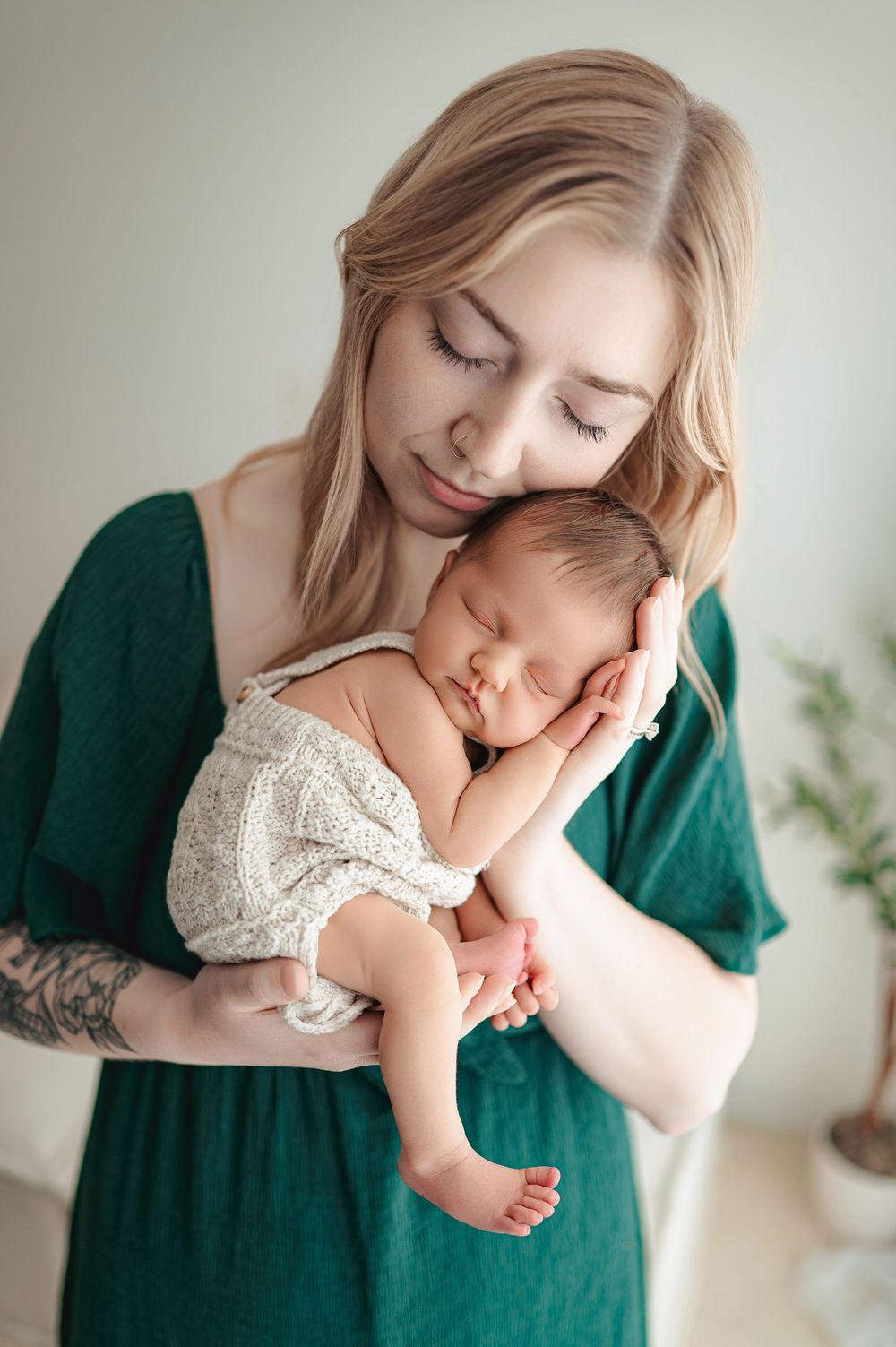 Tender moment as mother in emerald green dress cuddles sleeping newborn baby in white romper against light background.