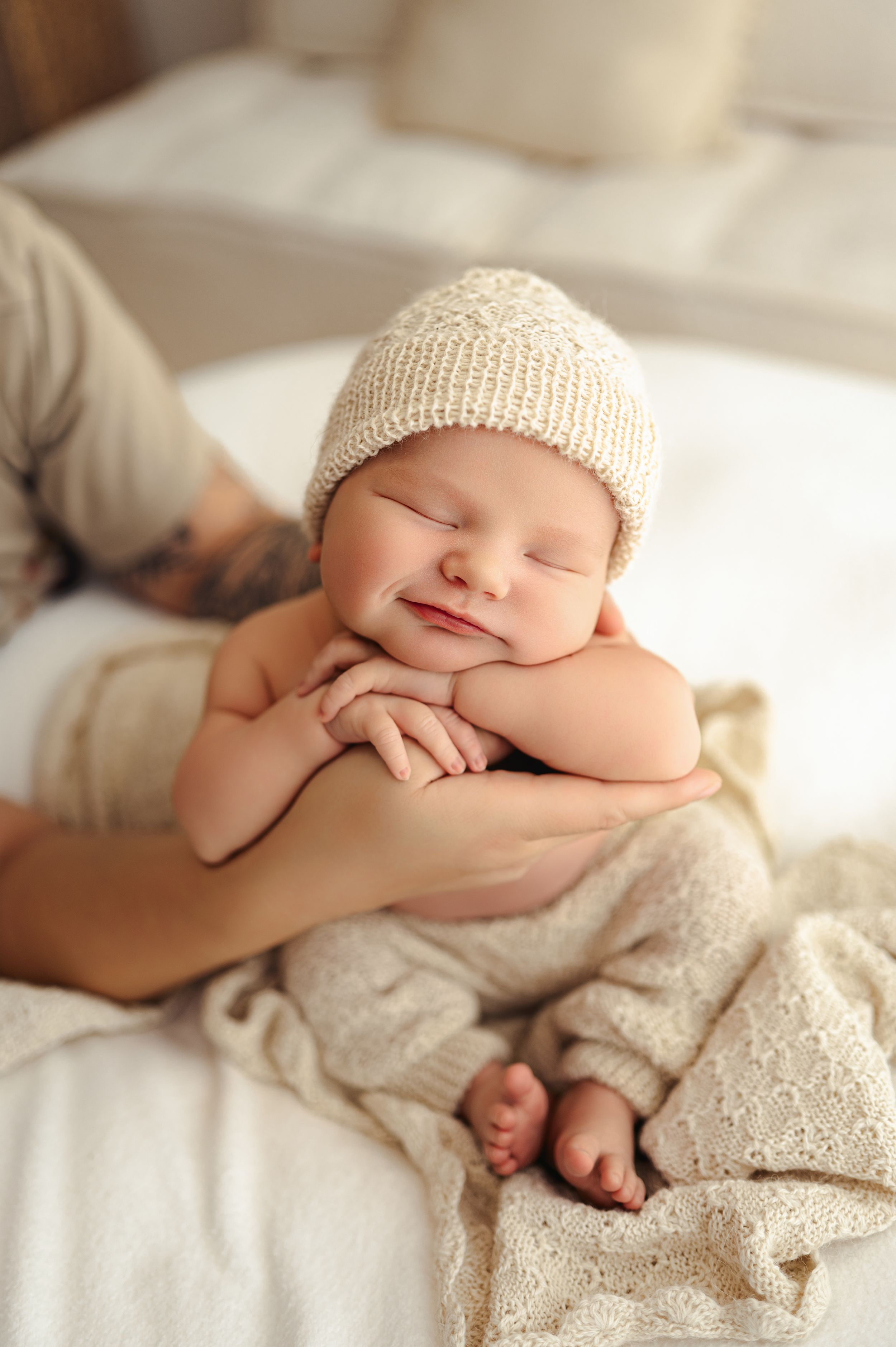 A sleeping newborn baby wrapped in a cream knit blanket and wearing a matching knit hat lies peacefully on white bedding.