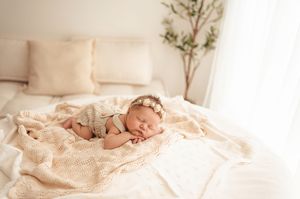 Sleeping newborn in light gray outfit rests peacefully on cream colored blanket near a potted plant in bright room.