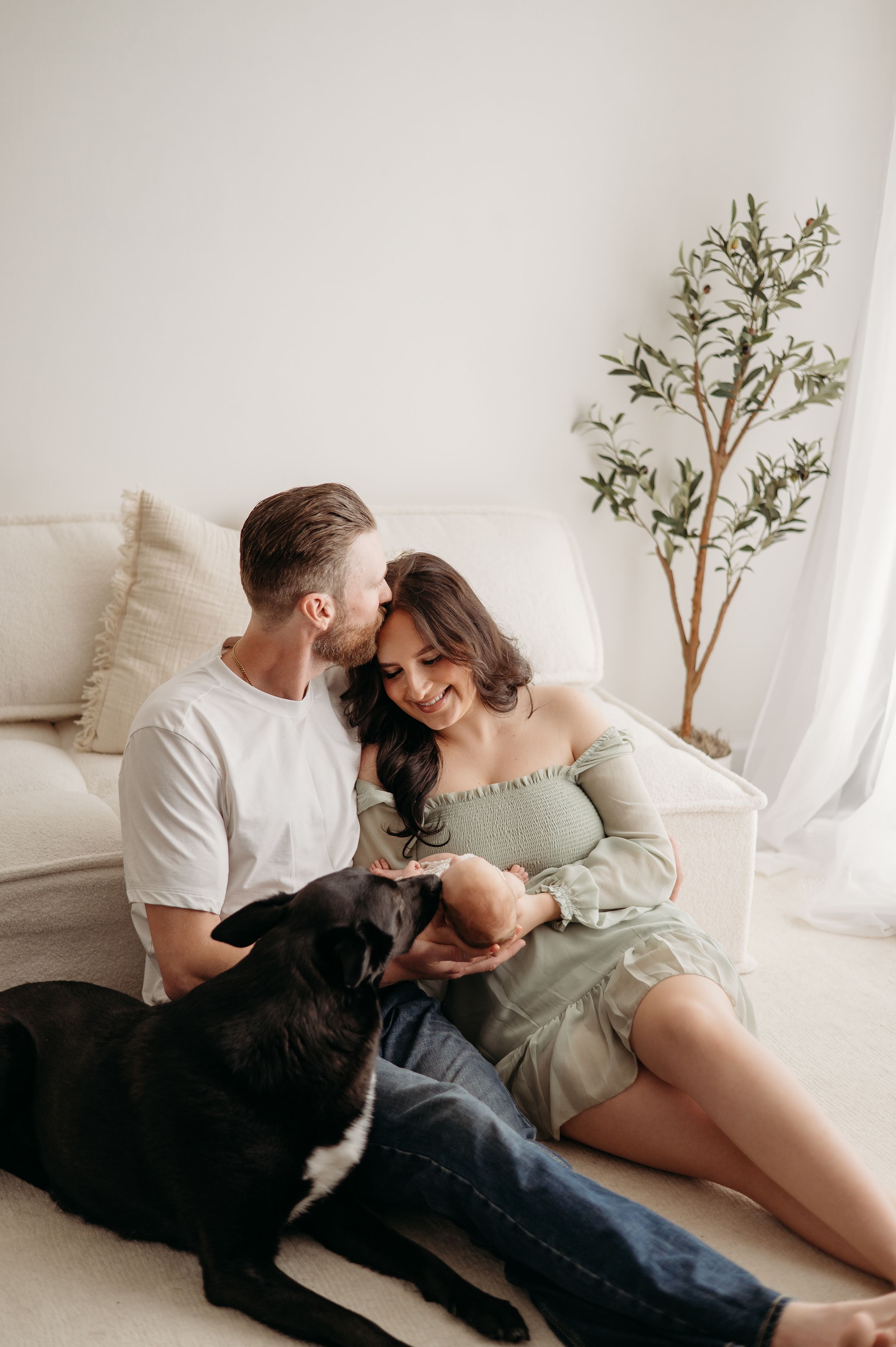 A couple relaxes together with their black dog in a bright minimalist room with white walls and a small potted plant.