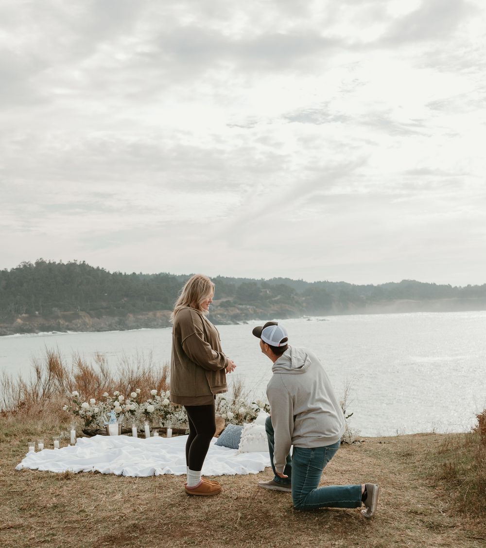 Two people share an intimate moment on a grassy overlook with a bay and mountains in the distance.