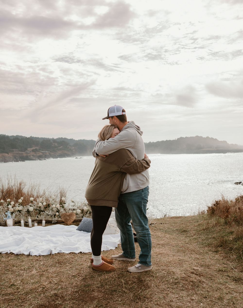 Two people embracing on a scenic overlook with a lake and mountains in the background.