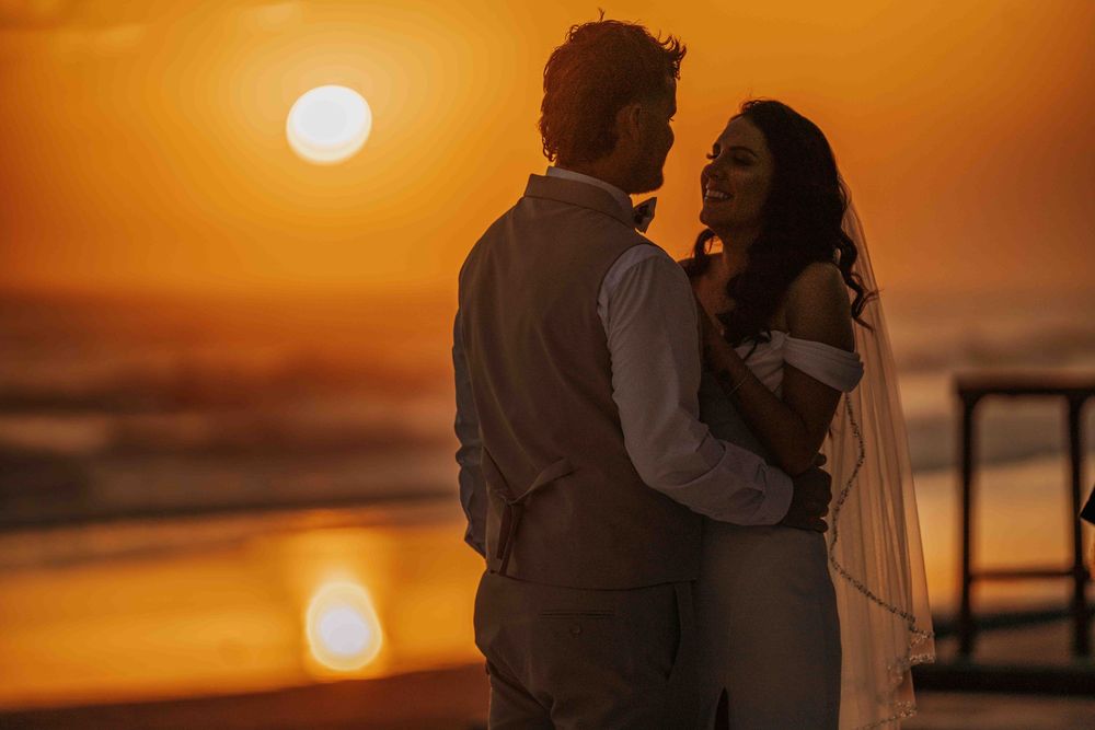 Two silhouettes embrace on a beach as the sun sets over the ocean, casting a warm orange glow across the horizon.