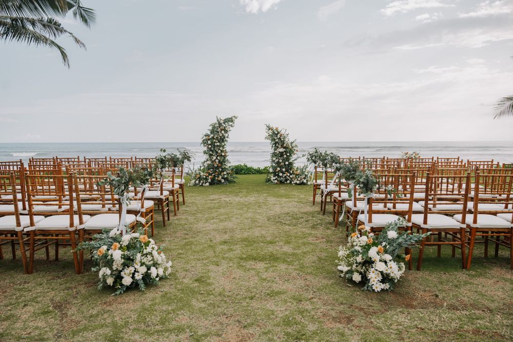 Rows of wooden chairs set up for an outdoor beach wedding ceremony with tropical palm trees and white floral arrangements with view of beach