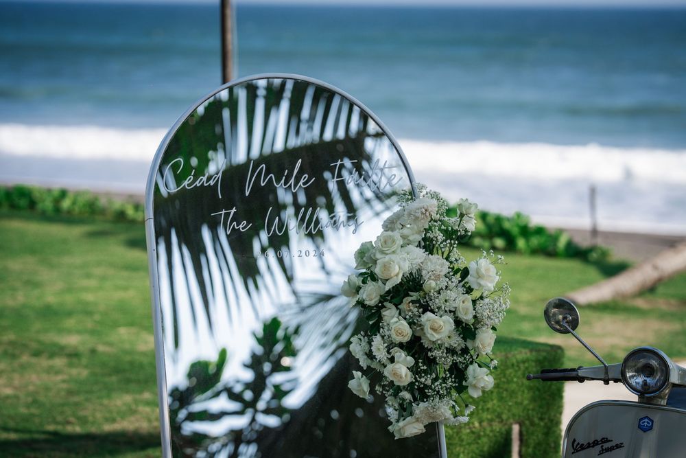 White floral arrangement and palm tree reflection in mirror at beachside wedding ceremony setup with ocean view.