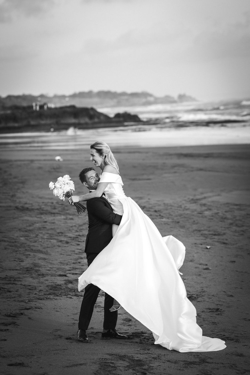A romantic black and white photo of a couple dancing playfully on a beach with headlands in the background.