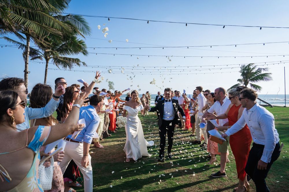 Wedding guests throw flower petals in the air during an outdoor beach celebration at sunset.