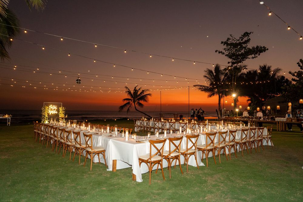 Elegant outdoor wedding reception setup with U-shaped table under string lights at sunset by the ocean.