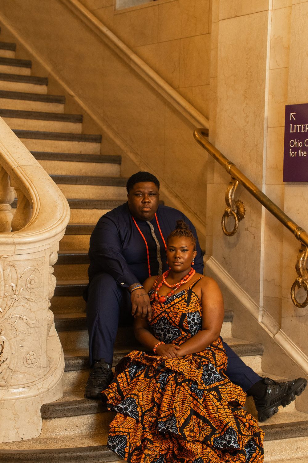 Couple in African attire sitting on marble stairs, woman in orange ankara dress, man in navy suit with red coral beads.