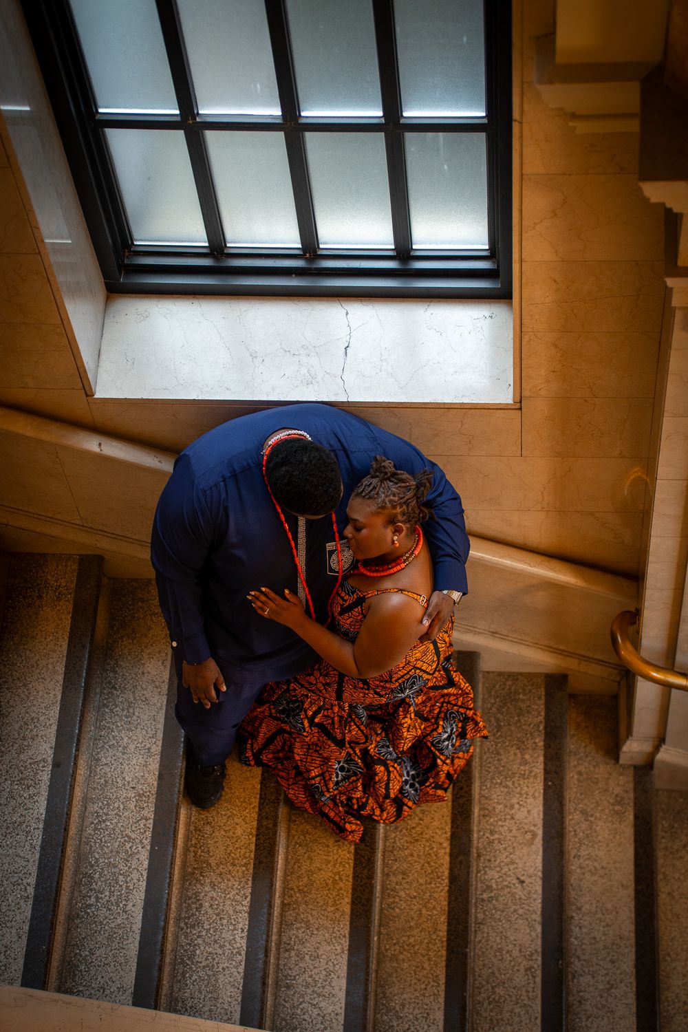 Aerial view of a couple in traditional African attire sharing a romantic moment on marble stairs near a window.