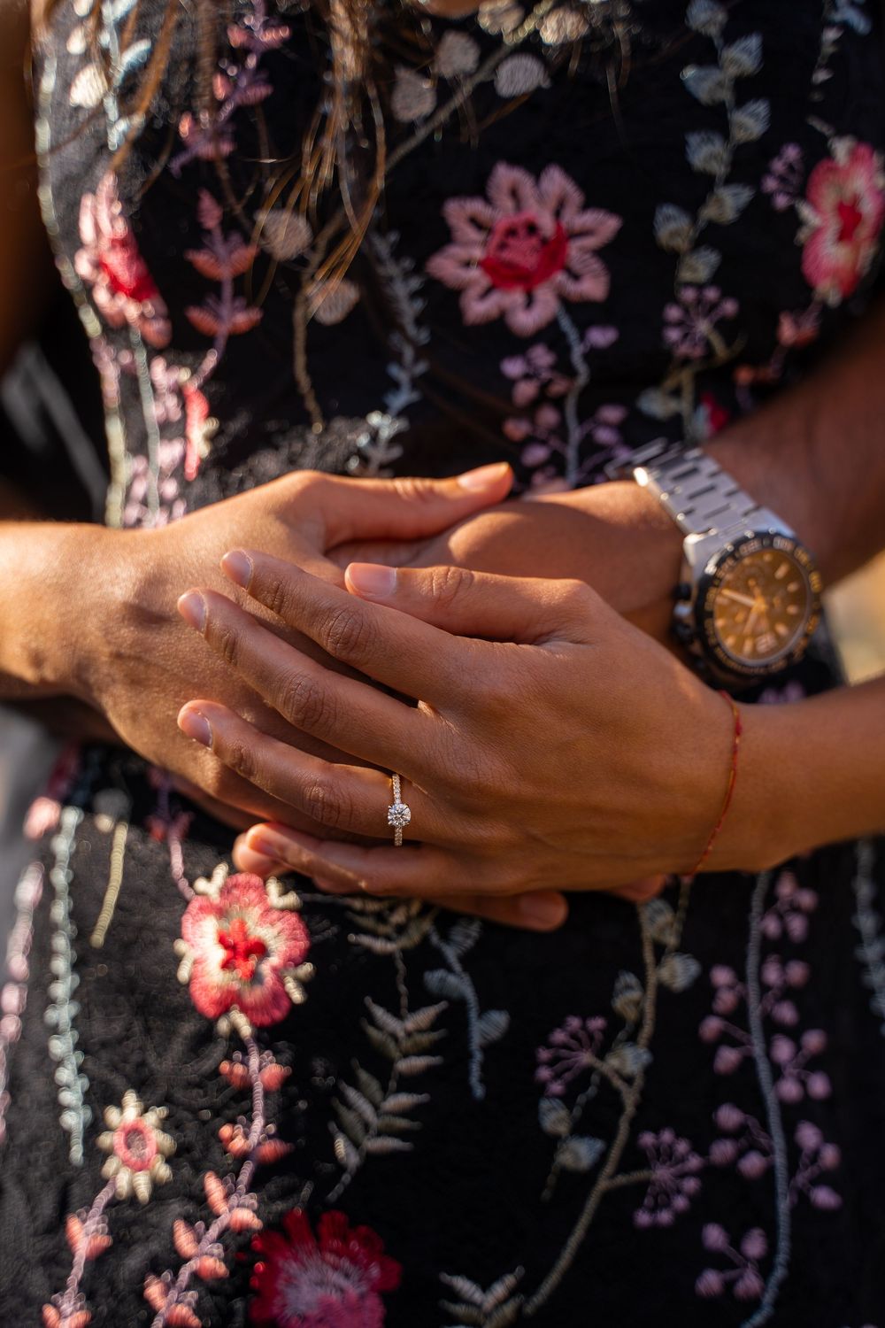 Close-up of couple holding hands showing engagement ring and watch, woman wearing floral embroidered black dress.
