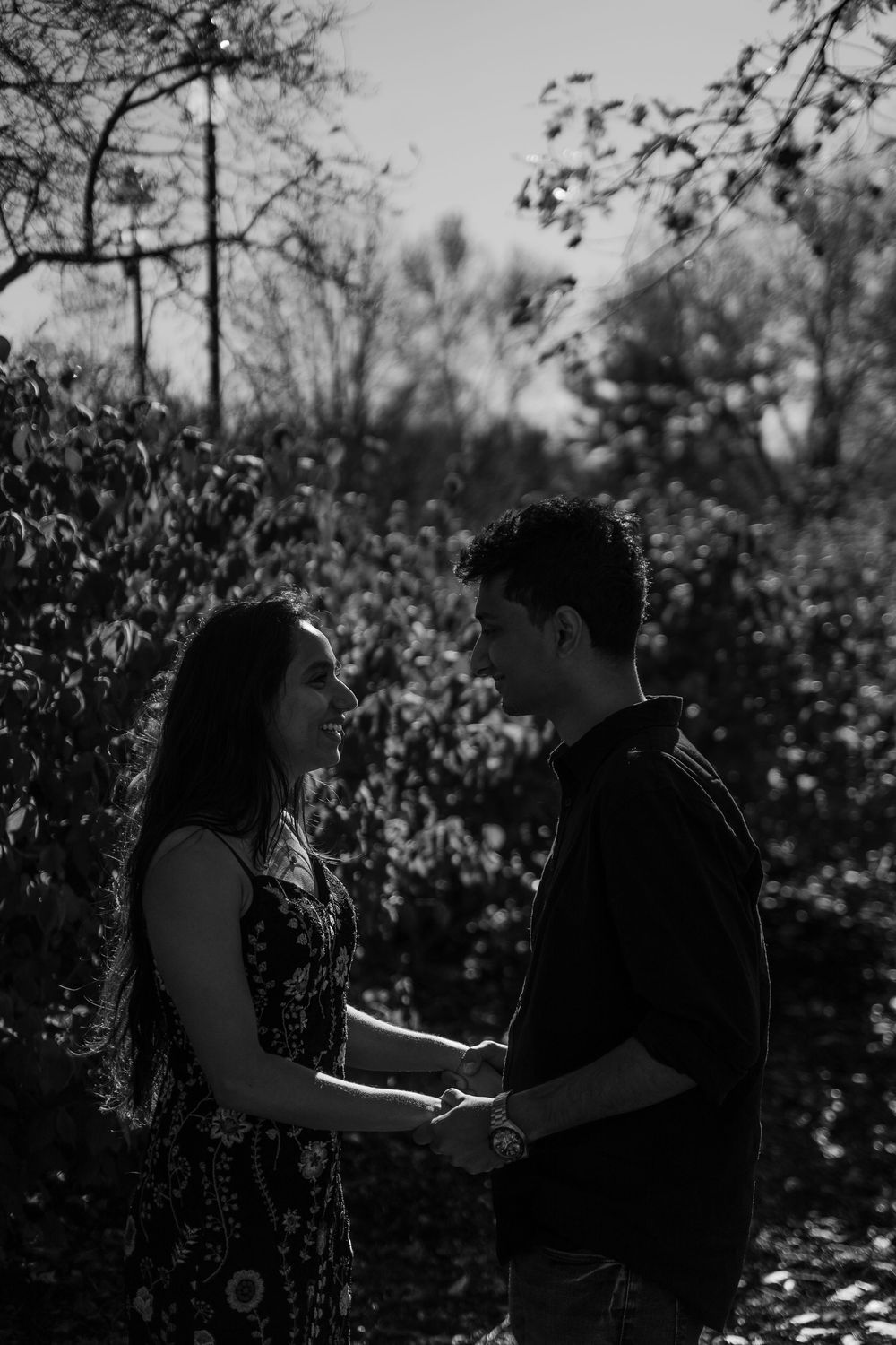 Black and white photo of a couple holding hands and gazing at each other in a lush outdoor setting.