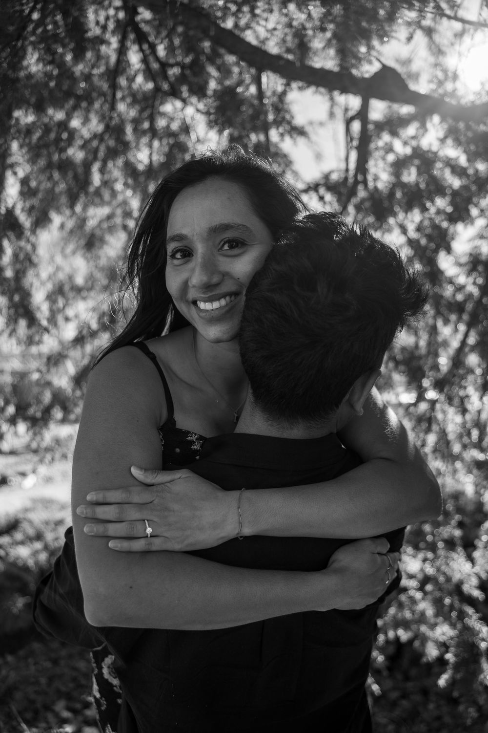 Black and white photo of a smiling woman hugging a man outdoors under trees.