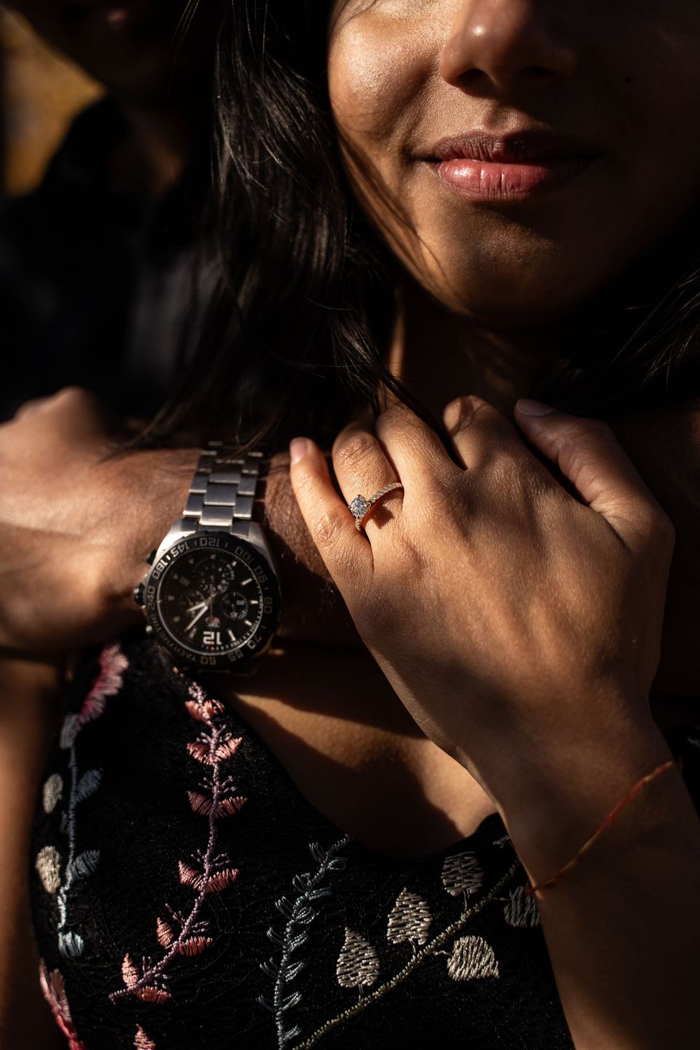 Close-up of woman in floral dress with mans hand wearing silver watch resting on her chest, ring visible.