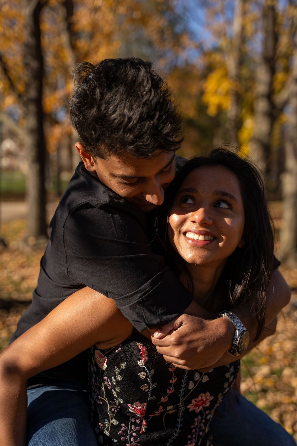 Couple embracing in autumn park, woman smiling up at man hugging her from behind among golden fall foliage.