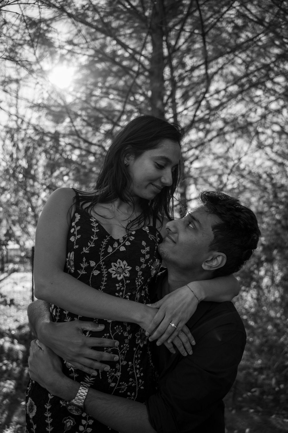 Black and white photo of a romantic couple embracing outdoors among trees, man looking up lovingly at woman in floral dress.