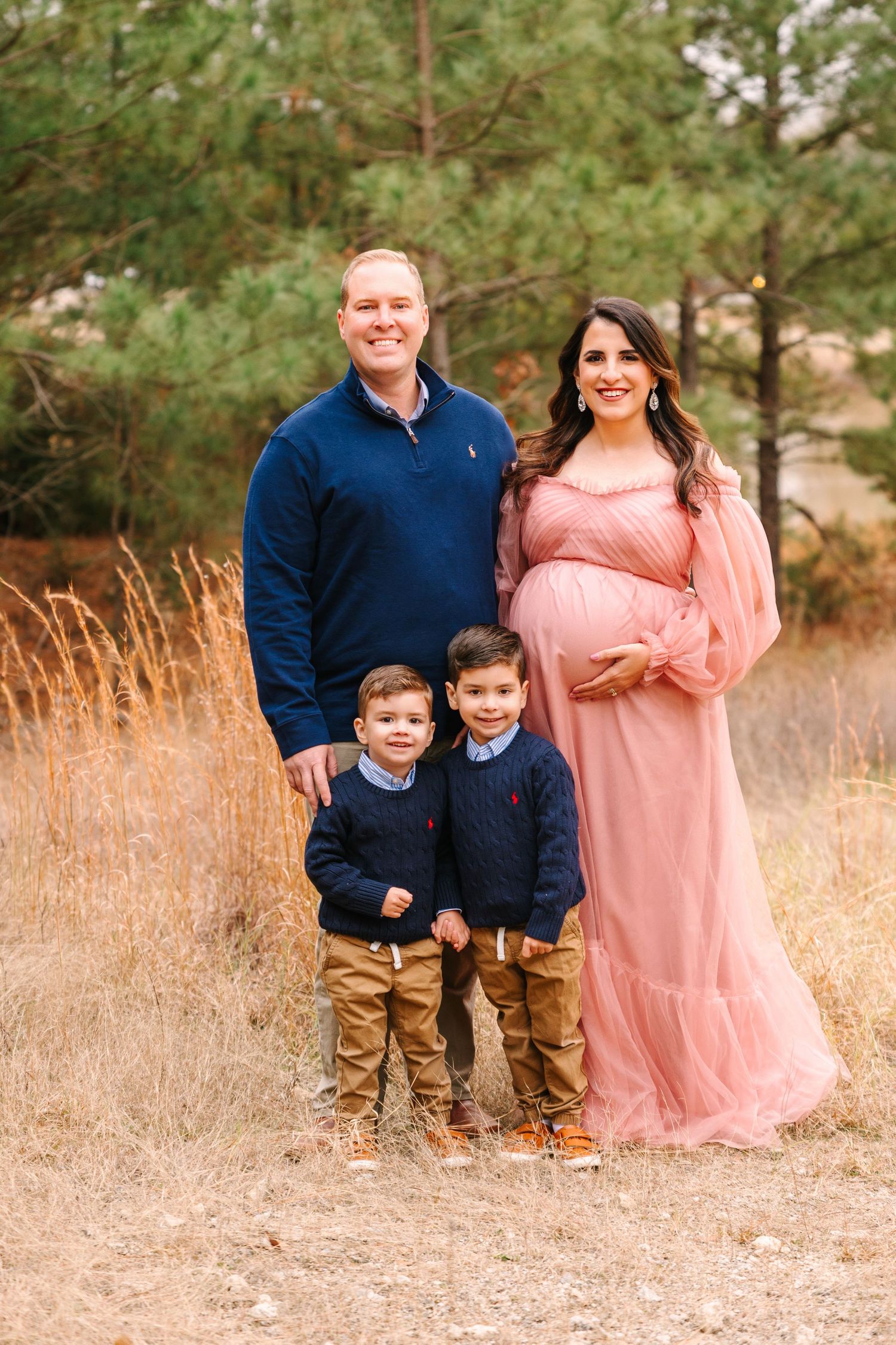 A family poses together in tall grass with pine trees in the background, wearing navy and pink coordinated outfits.