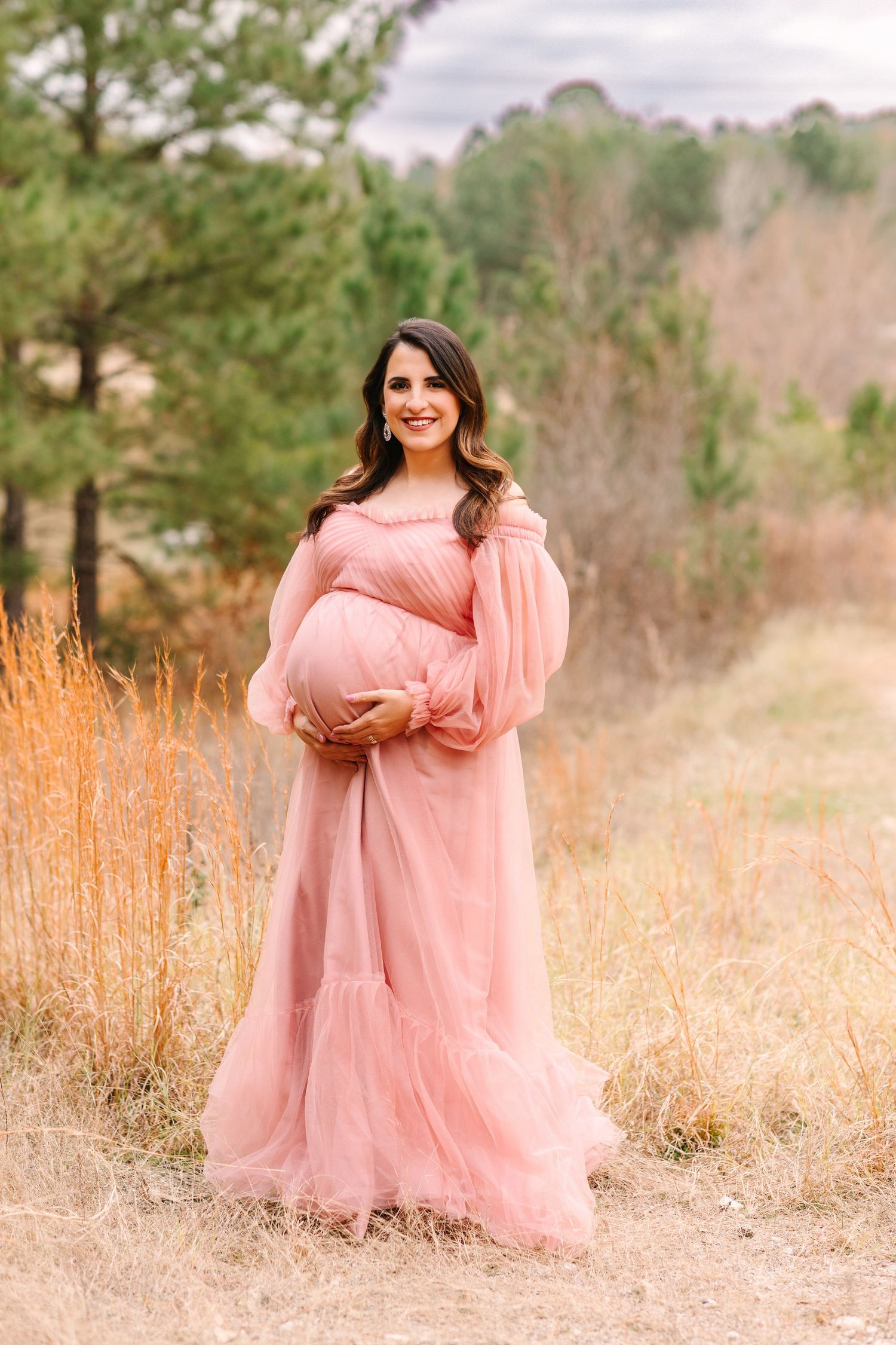 Pregnant woman in flowing pink gown poses in a field of tall grass during golden hour.